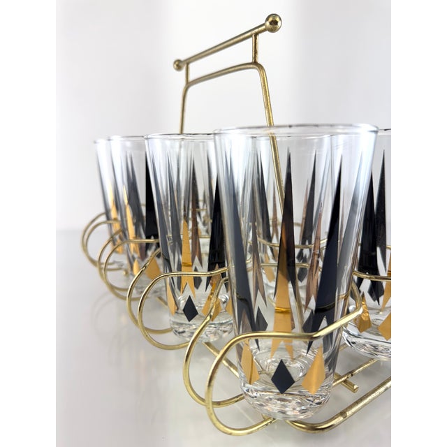 Mid-century modern bar set featuring eight highball glasses housed in a brass-toned carrier. Each glass is decorated with...