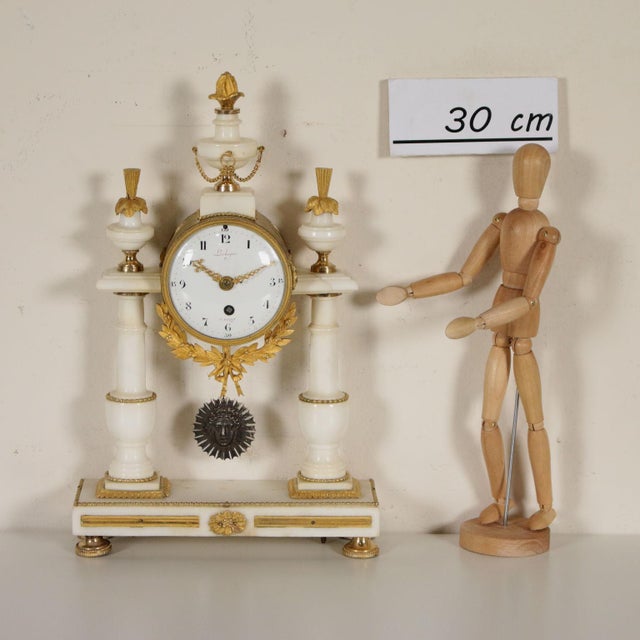 A table clock made of white marble with gilded bronze details. Rectangular basement with two turned columns and a...