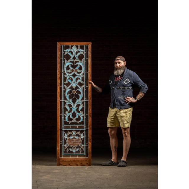 Tall oak-framed, leaded opalescent window in layered umber, teal, and ice-blue glasses with stylized foliate tracery and...