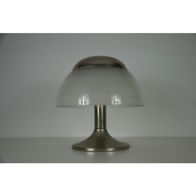 The lamp features a cylindrical base in metal, typically finished in brushed steel or chromed brass, supporting a domed...