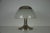 The lamp features a cylindrical base in metal, typically finished in brushed steel or chromed brass, supporting a domed...