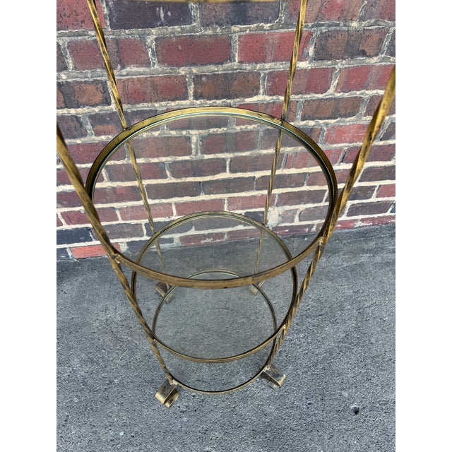 Vintage brass tone iron display shelf with four glass shelves. The frame features scroll details at the top and feet,...
