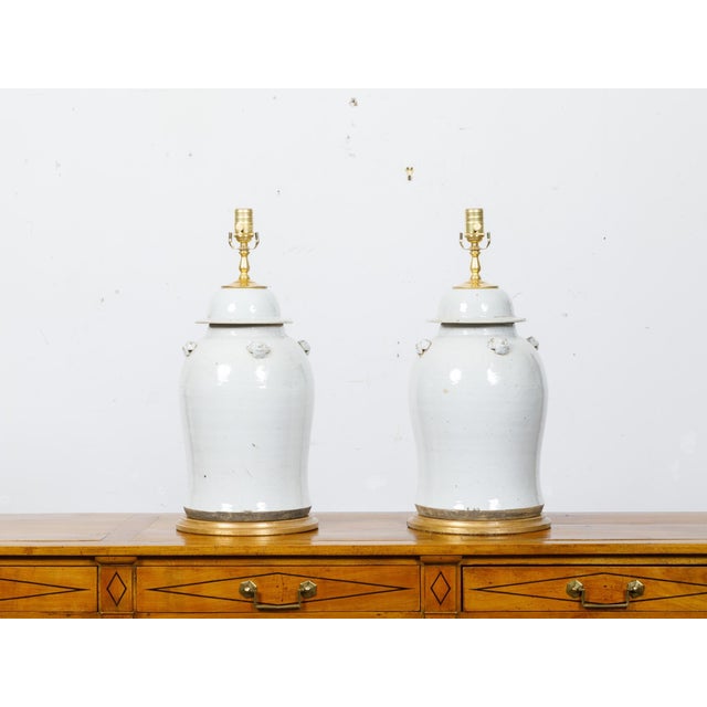 A pair of white porcelain lidded urns table lamps with single lights on circular gilded bases, wired for the USA. This...