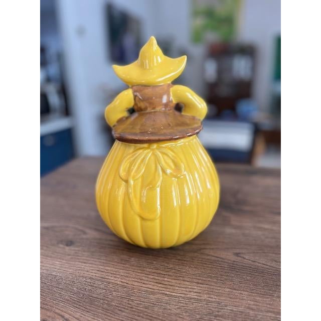 The jar is made of ceramic with a vibrant yellow glaze. It features a Dutch girl design with braids, wearing a yellow...