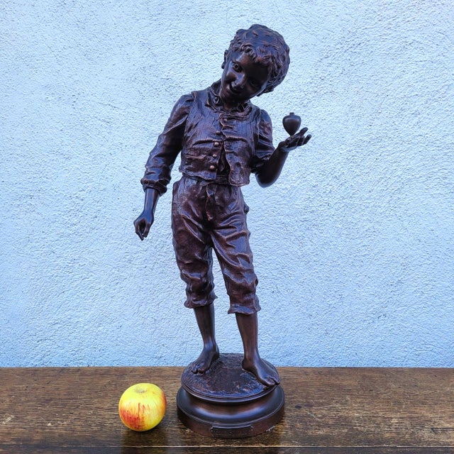 Beautiful bronze sculpture with a brown patina, depicting a young boy holding a spinning top. The bronze is titled "She's...