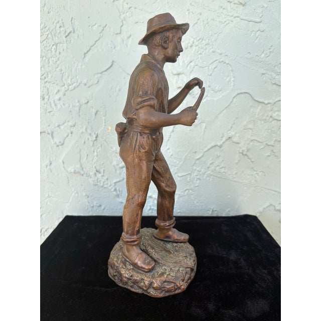 This antique figurative sculpture depicts a young working boy, captured mid-gesture and holding a hand tool, rendered with...