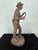 This antique figurative sculpture depicts a young working boy, captured mid-gesture and holding a hand tool, rendered with...
