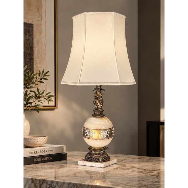 Ornate table lamp with a cream/ivory glass globe body, a gold-tone floral band, a cherub detail and an intricately cast...