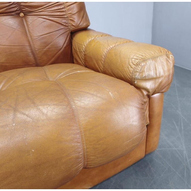 The sofa is upholstered in cognac leather and features deep tufted cushions, generous padding and sculptural armrests that...