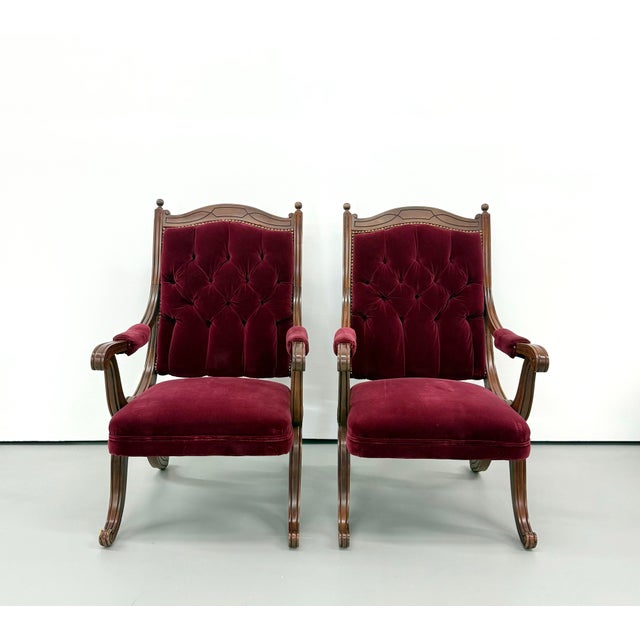 A dramatic pair of vintage high-back armchairs featuring deep burgundy velvet upholstery with generous button tufting...