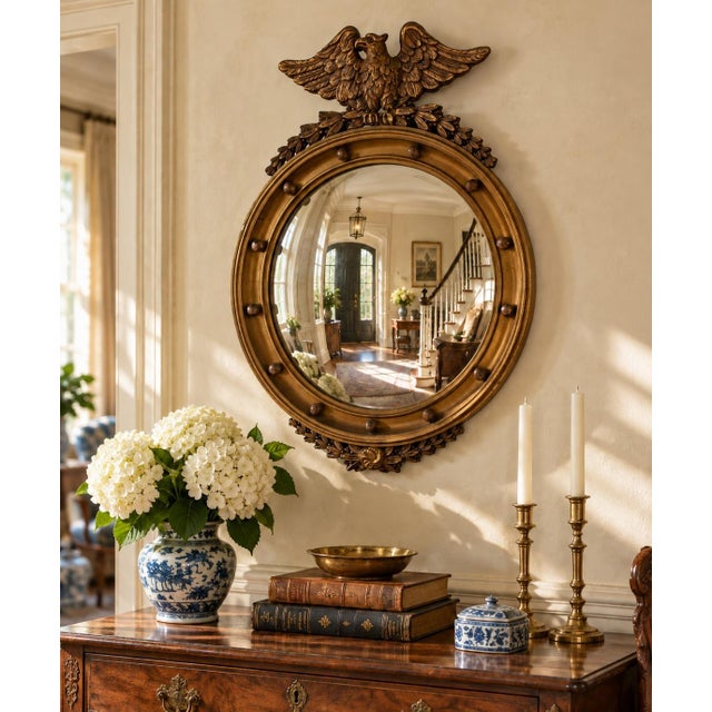 A Federal-style giltwood convex mirror crowned with a carved eagle, its wings outstretched above a laurel motif—an...