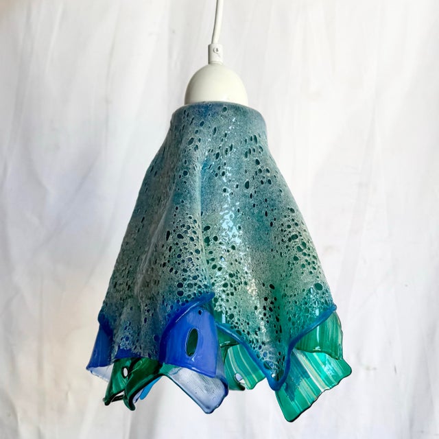 A striking hand-blown glass pendant lamp featuring an organic, sculptural shade with fluid ruffled edges reminiscent of a...