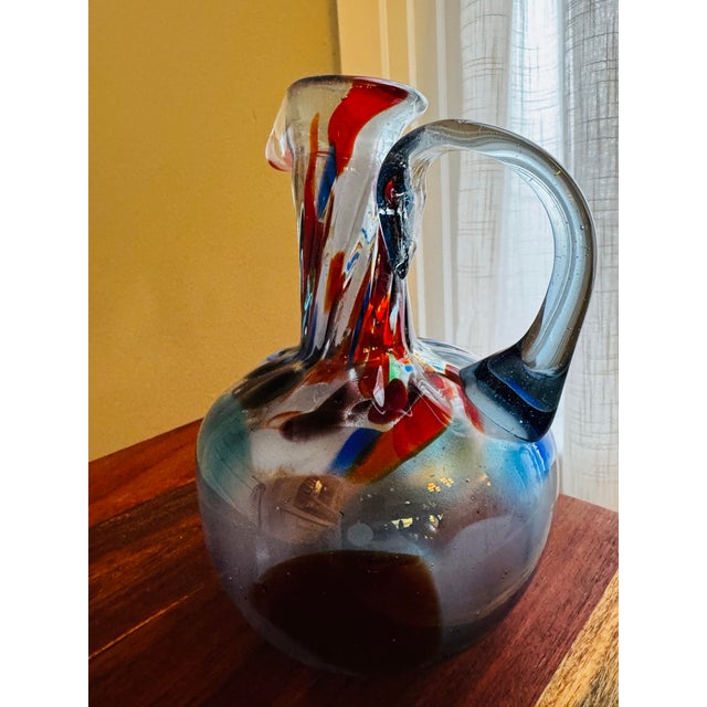 This sculptural hand-blown glass pitcher features a striking abstract composition of blue, amber, red, and cobalt...