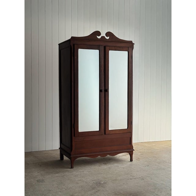 Antique mahogany wardrobe cabinet with mirrored doors and a shaped scroll crest, the surface showing warm red-brown tones...