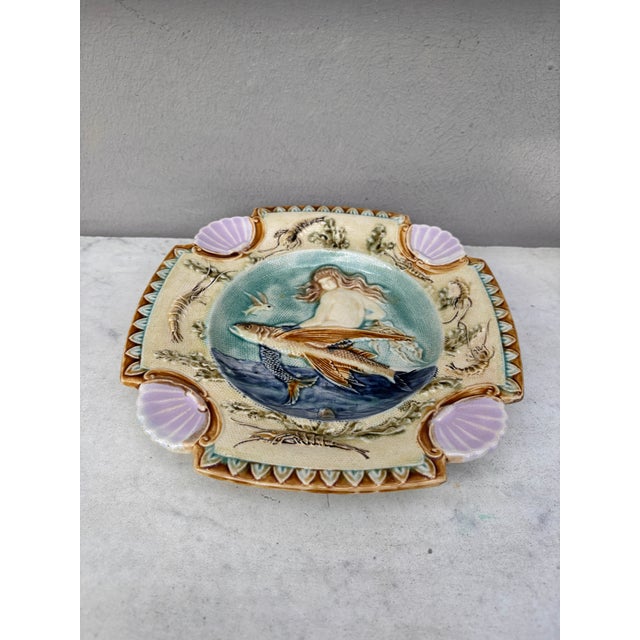 Square Majolica mermaid plate with shells on the corners, the mermaid sit on a flying fish, the border is decorated with...