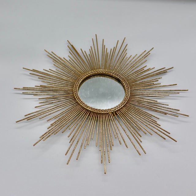 A classic sunburst/starburst wall mirror featuring radiating metal rays in a warm gold-toned finish, centered around a...