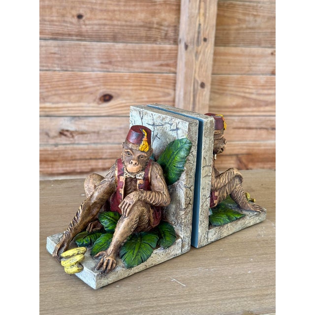 A whimsical pair of cast resin bookends featuring monkeys dressed in red vests and fez hats, perched on jungle leaves and...