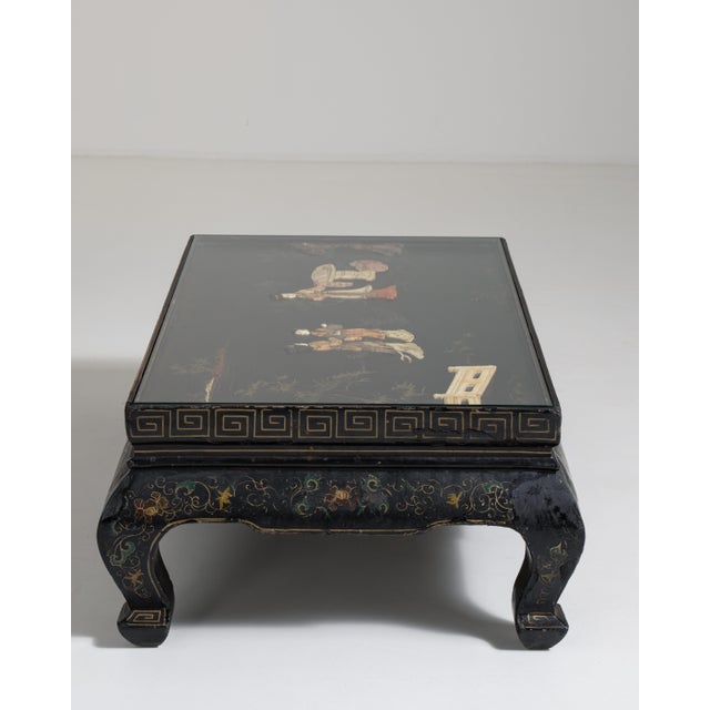 A decorative coffee table inspired by Chinoiserie motifs, featuring a glass top that highlights the detailed base. The...