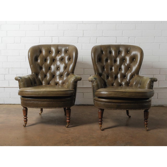 A handsome pair of vintage tufted leather club chairs featuring deep button-tufted backs, rolled arms, and elegant turned...