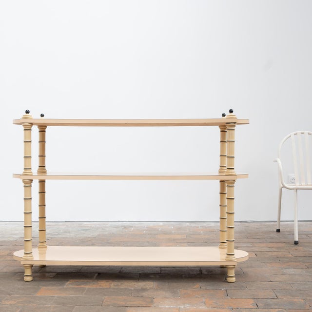 A 1970s display console in cream-beige lacquered wood, featuring three open shelves. Its decorative structure rests on...