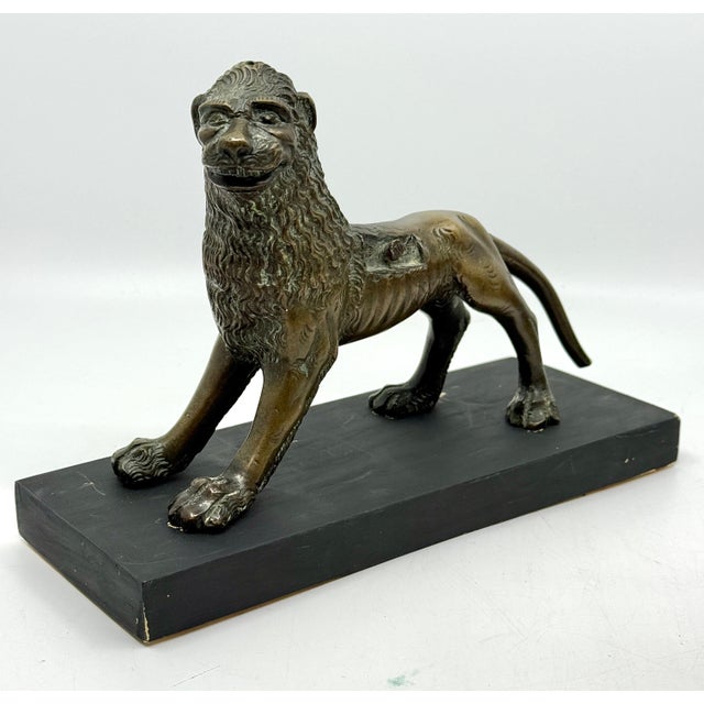 A cast bronze lion figure mounted on a black rectangular base, depicted in a dynamic stance with stylized detailing,...