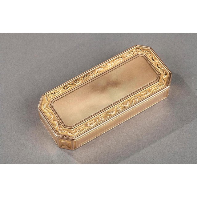 Rectangular gold box with cut sides. The hinged gold lid is adorned with concentric lines framed by a floral frieze of...