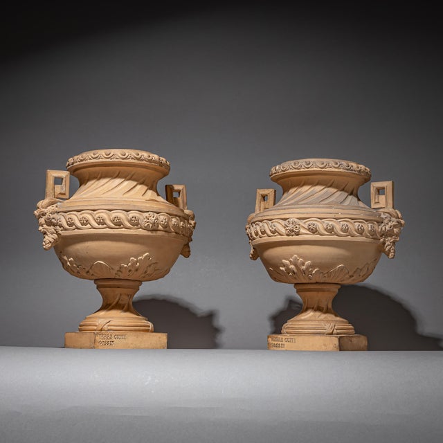 Pair of terracotta amphorae on square plinths, decorated with acanthus leaves and wave-band motifs. The side handles are...