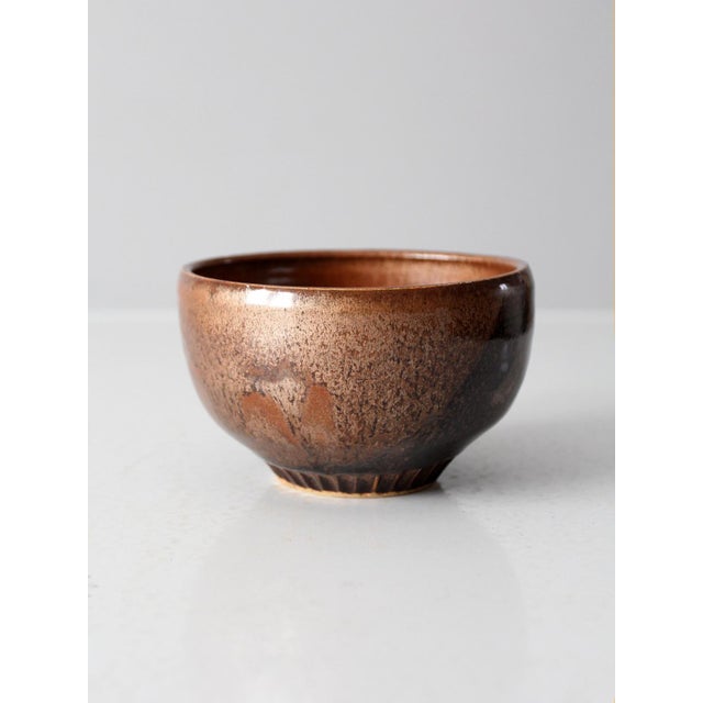 A vintage handmade studio pottery bowl with a richly mottled glaze in warm copper brown, umber, and near-black tones. The...