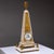 Obelisk-shaped marble clock with fire-gilt bronze appliqués, relief scene, and a surmounting armillary sphere. The clock...