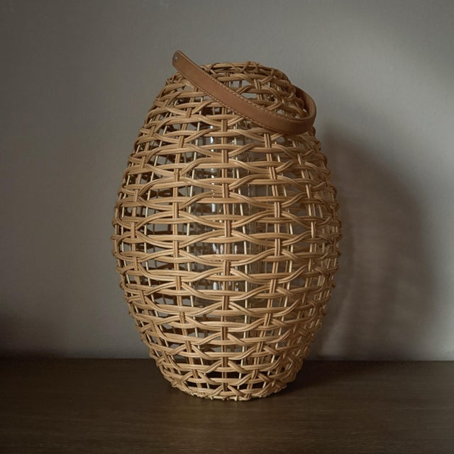 A sculptural oval lantern by Two’s Company, crafted in handwoven natural rattan and finished with a warm faux leather...