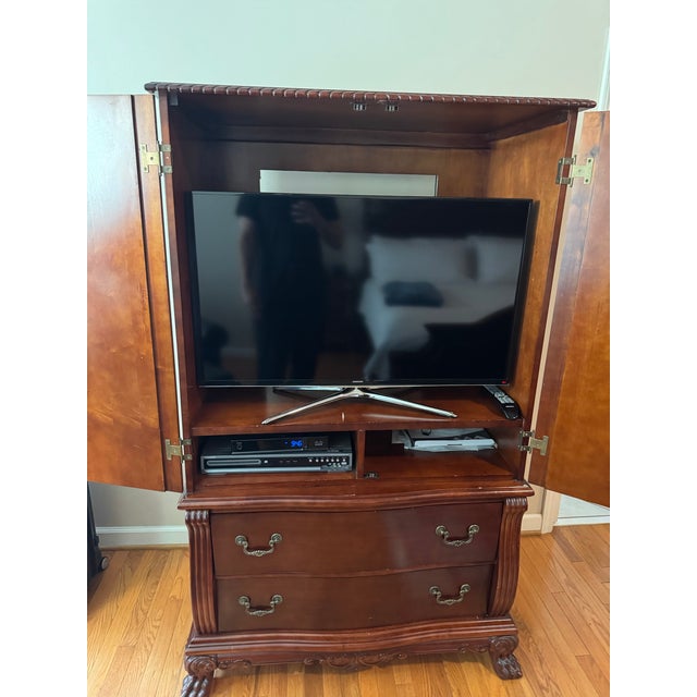 A traditional cherry-finish armoire from a four-piece carved bedroom suite. Two arched raised-panel doors open to a shelf...
