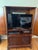 A traditional cherry-finish armoire from a four-piece carved bedroom suite. Two arched raised-panel doors open to a shelf...