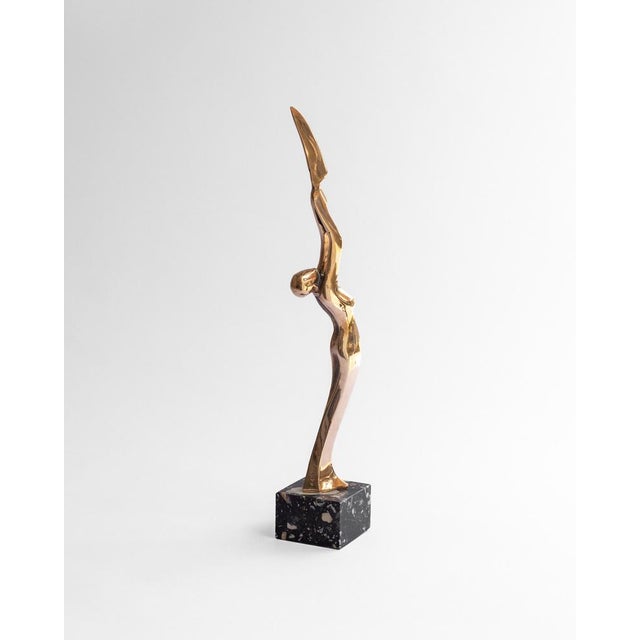 A polished bronze sculpture depicting an abstracted female figure in a graceful, arched pose, arms raised and merging into...