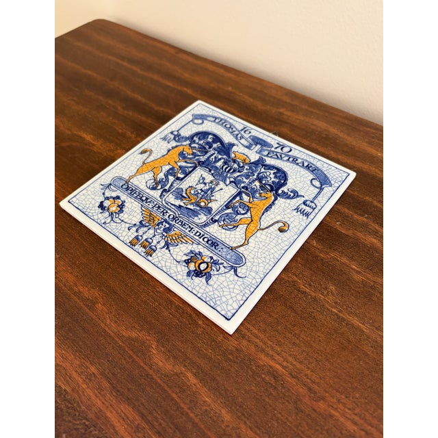 A classic Dutch Delft-style hand painted blue and white porcelain tile featuring an intricately rendered heraldic coat of...