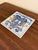 A classic Dutch Delft-style hand painted blue and white porcelain tile featuring an intricately rendered heraldic coat of...