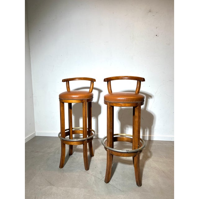 A elegant pair of vintage bar stools combining warm solid wood construction with tan leather upholstery and a brass-toned...