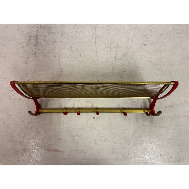 A mid-century wall-mounted rack combining a coat rail and upper shelf, executed in brass with red-lacquered supports. The...