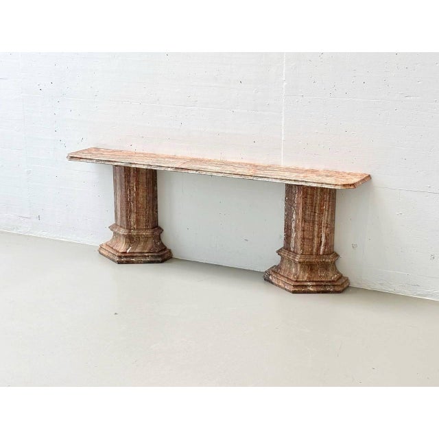 The piece of furniture shown is a striking console table made of reddish-brown marble with vibrant veining and a polished...