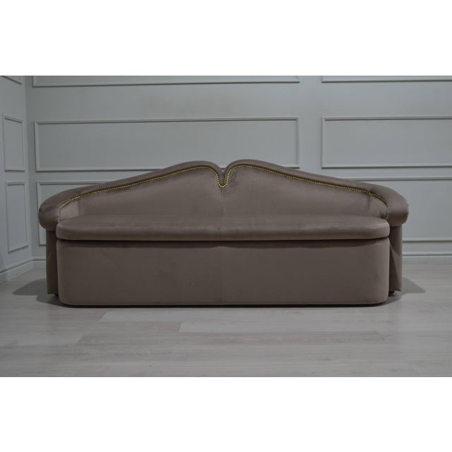 An elegant vintage-inspired sofa upholstered in warm taupe fabric, featuring a distinctive sculpted backrest with a...