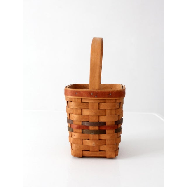 A 1992 Longaberger handwoven basket in warm honey-toned wood with woven red and green accent bands along the rim. The tall...