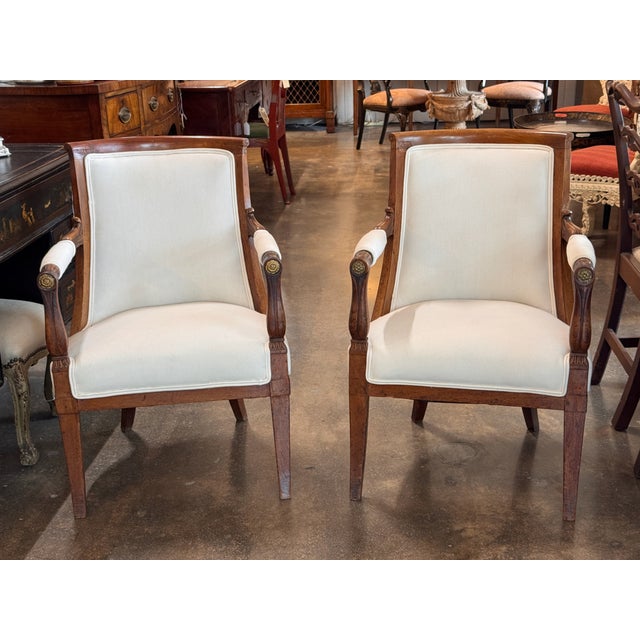 Pair of French Empire–style armchairs with curved backs, scroll arms, and carved wood frames. Upholstered seats and backs...