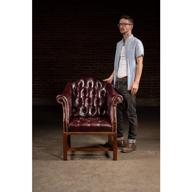 A stately mid-century leather club chair upholstered in deep burgundy tufted leather with brass nailhead trim and a solid...