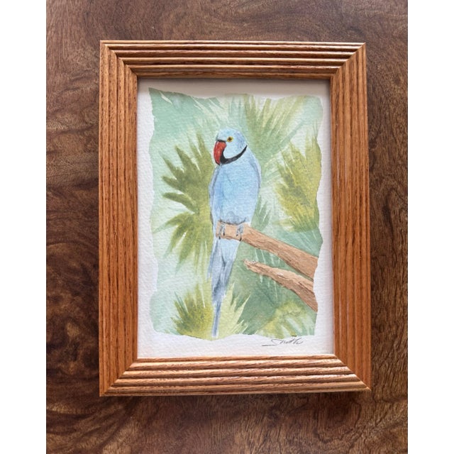 A delicate blue parakeet rests peacefully on a branch, surrounded by leafy green foliage. The soft watercolor technique...