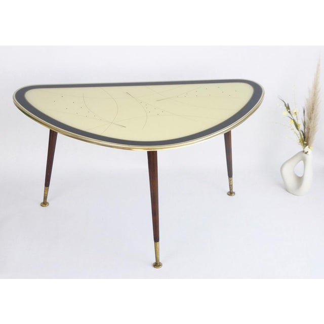 A large kidney shaped glass formica table with a brass rim standing on wooden legs. It has a nice abstract design. This...