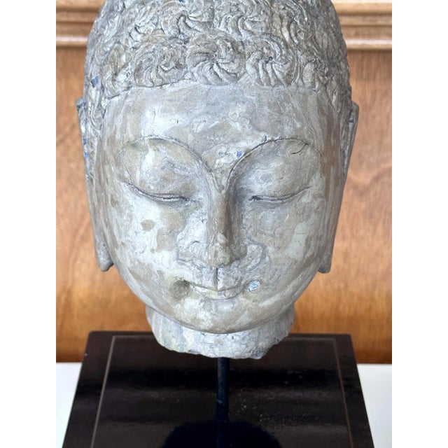 Carved out of a single block of grayish brown stone (likely granite type), this Buddha head represents Northern Qi style...