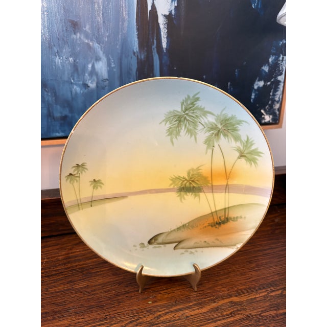 Featuring a sunset landscape with palm trees in soft pastel tones, finished with a gilt rim. Backstamped “Hand Painted...