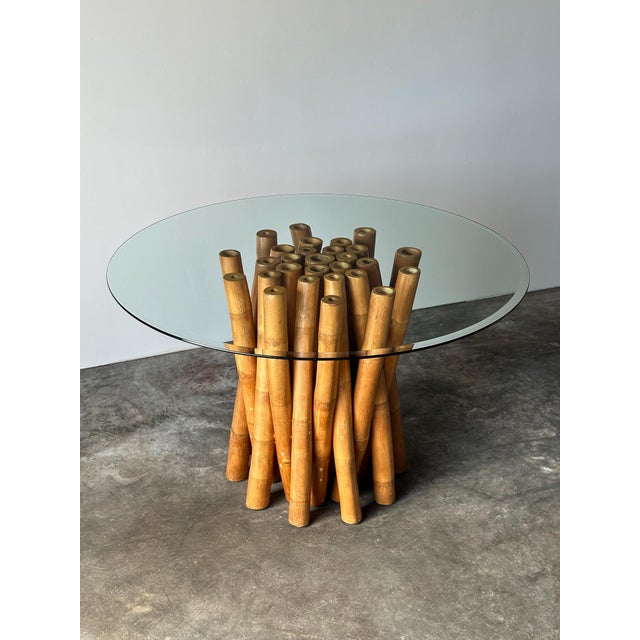 A round glass-top dining table raised on a sculptural base of dozens of natural bamboo poles, bundled and splayed outward...