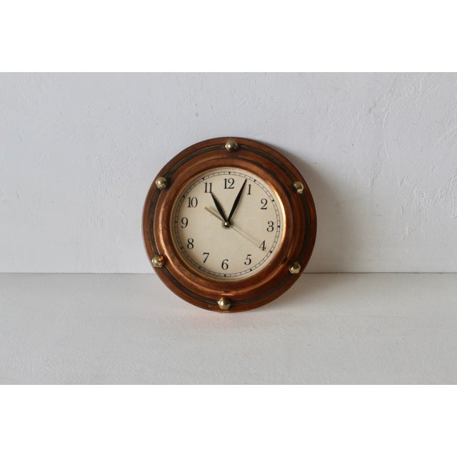 Vintage nautical porthole-style wall clock with a warm, copper frame, brass-tone bolt accents, and a classic off-white...