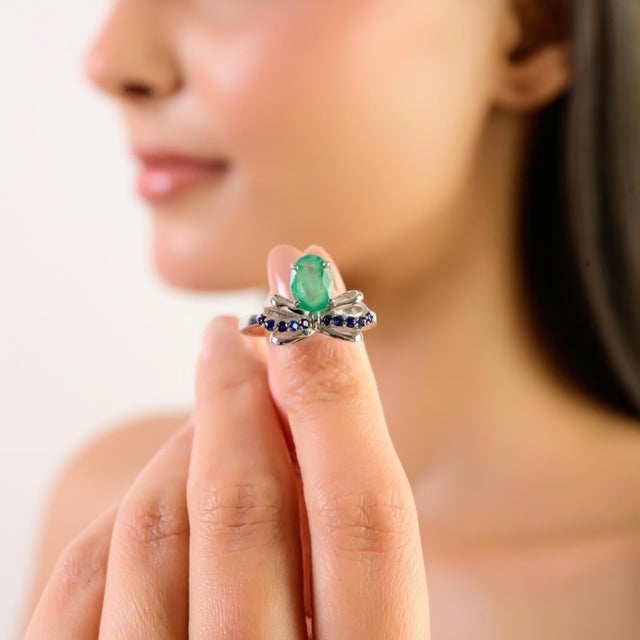 This modernist sterling silver ring features an elegant bow motif centered with an 8x6 mm oval emerald. Sparkling round...