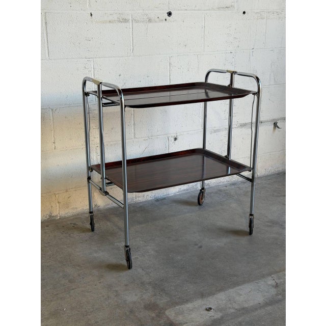This versatile rolling utility cart features a sleek folding chrome frame and rich rosewood-patterned laminate trays. The...